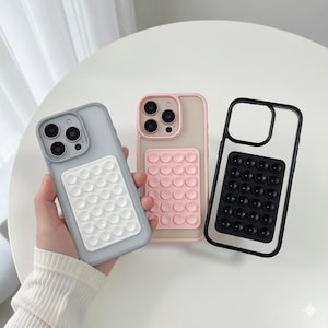 Puede incluir: Tres fundas para teléfono con diseño de burbujas. Una funda es gris claro con un panel de burbujas blanco, otra es rosa con un panel de burbujas rosa y la tercera es negra con un panel de burbujas negro. Las fundas están sobre una superficie blanca.