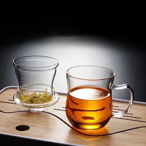 May include: A clear glass tea cup with a handle, filled with amber liquid, sits next to a glass infuser with tea leaves. The tea set is on a bamboo tray, against a dark background.