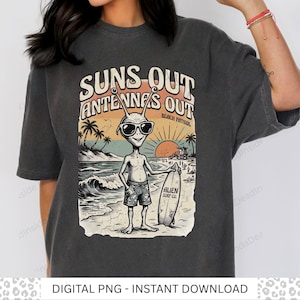 May include: A charcoal gray t-shirt featuring a graphic print of an alien wearing sunglasses and patterned swim trunks while holding a surfboard on a beach. The text reads "SUNS OUT ANTENNAS OUT" and "BEACH PATROL" with "ALIEN SURF CO." on the board.