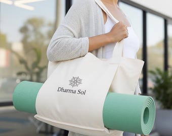 May include: A cream-colored tote bag with the text "Dharma Sol" and a logo, holding a rolled-up green yoga mat. The bag is carried over the shoulder. The yoga mat is approximately 68 inches long.