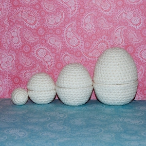 May include: Four white crocheted eggs of different sizes, arranged from smallest to largest, on a blue background.