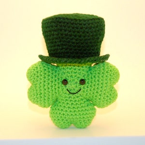 Pode incluir: Um trevo de quatro folhas de crochê verde usando um chapéu de coco de crochê verde com uma faixa amarela. O trevo tem um rosto sorridente com olhos pretos.
