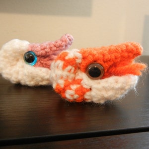 May include: Two crocheted amigurumi toys shaped like sea creatures. One is pink and white with a blue eye, the other is orange and white with a brown eye.