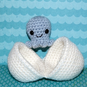 May include: A crocheted light blue octopus with black eyes and a smiling face sits above a white crocheted egg. The egg is open, revealing the octopus. The background is a blue pattern with white waves.