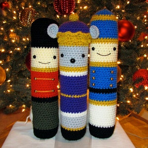 May include: Three crocheted nutcracker ornaments, each with a different design. The first is a red and black nutcracker, the second is a purple and white nutcracker, and the third is a blue and white nutcracker. All three ornaments have a smiling face and are standing on a white surface.