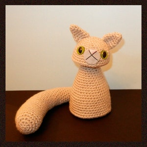 Puede incluir: Un juguete de gato de crochet beige con grandes ojos verdes y una nariz rosa. El gato tiene una cola larga y rizada.