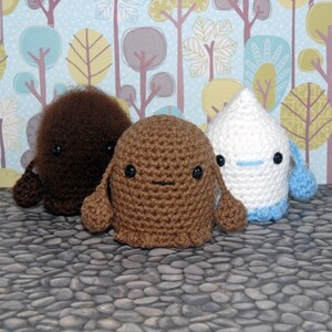 May include: Three crocheted stuffed toys, one brown, one white, and one dark brown, with embroidered eyes and mouths. The toys are sitting on a gray and white stone surface.