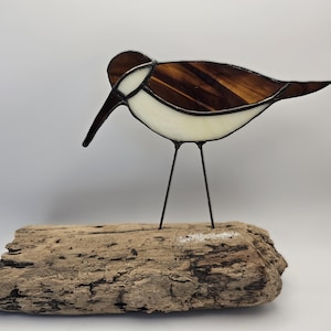 Puede incluir: Una escultura de pájaro de vidrieras con cuerpo marrón y blanco, pico negro y finas patas negras. El pájaro está posado sobre un trozo de madera flotante. El vidrio tiene una apariencia texturizada.
