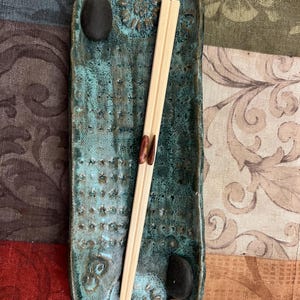 May include: A rectangular, turquoise ceramic chopstick rest with a textured surface and floral designs. Two small black stones and a pair of wooden chopsticks rest on the tray. The tray is approximately 8 inches long.