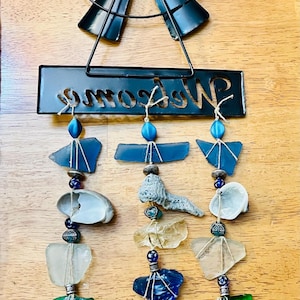 May include: A rustic wind chime featuring a black metal windmill design at the top, with a rectangular sign that reads "Welcome". The chime includes sea glass, shells, and beads in shades of blue, green, and white, suspended by twine.