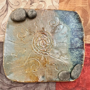 May include: A square ceramic plate with a textured surface and a spiral design in the center. The plate has a blue and brown color scheme, with small stones on the corners. Decorative pottery.