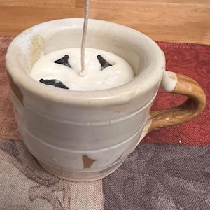 May include: A handmade ceramic mug candle with a cream-colored wax and a wick. The candle has three black accents and a brown handle. The mug is off-white with brown accents and horizontal lines.