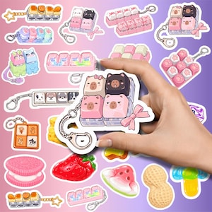 Puede incluir: Una colección de pegatinas con varios llaveros de teclas de teclado mecánico con caras de animales, huellas y diseños de comida, incluyendo una galleta sándwich rosa, una fresa roja, una rodaja de sandía y un cacahuete sobre un fondo degradado.