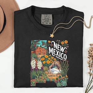 May include: Black crew neck t-shirt with a colorful graphic of New Mexico's landscape, including the text "NEW MEXICO" and "SANTA FE". A gold star pendant necklace rests on the shirt. The shirt has a Comfort Colors tag.