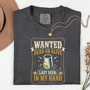 May include: Dark gray t-shirt with the text "WANTED DEAD OR ALIVE" and a graphic of a socket wrench. The text "LAST SEEN: IN MY HAND" is also printed on the shirt. A gold necklace with a star pendant is on the shirt.