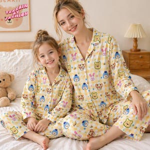 May include: Matching yellow pajamas with cartoon character prints. The pajamas feature a button-down shirt and pants. The image shows a mother and child wearing the pajamas. The background includes a teddy bear and a framed picture.