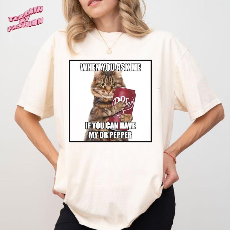 May include: Cream-colored t-shirt featuring a graphic of a cat holding a Dr. Pepper can. The image includes the text: "WHEN YOU ASK ME IF YOU CAN HAVE MY DR PEPPER." The shirt has short sleeves and a crew neck.