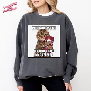 May include: Dark grey sweatshirt featuring a humorous graphic of a cat holding a Dr. Pepper can. The text on the graphic reads "WHEN YOU ASK ME IF YOU CAN HAVE MY DR PEPPER". The sweatshirt has a crew neck and long sleeves.