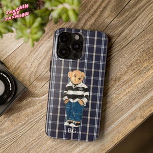 May include: A navy blue and white plaid phone case featuring a cartoon bear wearing a striped shirt and jeans. The case has the initials "BG" at the bottom and the text "TERRAIN BY FASHION" at the top.