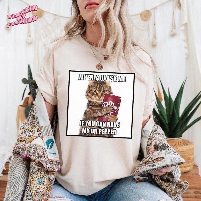 May include: Cream-colored t-shirt featuring a graphic of a cat holding a Dr Pepper can. The text on the graphic reads "WHEN YOU ASK ME IF YOU CAN HAVE MY DR PEPPER". The shirt has short sleeves and a relaxed fit.