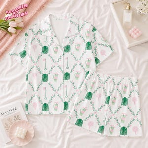 May include: White pajama set with a golf-themed pattern. The set includes a short-sleeved button-down shirt and matching shorts. The pattern features green golf course elements, pink flowers, and green vines. The text "TERRAIN BY FASHION" is visible.