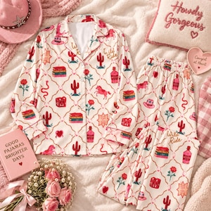 May include: A cream-colored pajama set featuring a repeating pattern of red cacti, pink horses, roses, snakes, cowboy hats, and books. The long-sleeved button-down top and matching pants include the text Wild Ride printed in a cursive script.