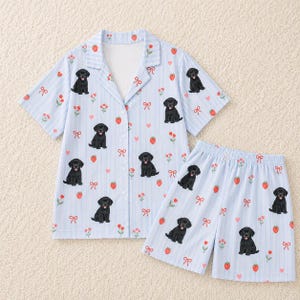 May include: A light blue and white striped pajama set consisting of a short-sleeved button-down shirt and matching shorts. The fabric features a repeating pattern of black Labrador puppies, red strawberries, small red flowers, and red bows.