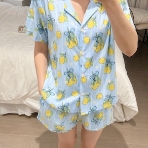 May include: A light blue pajama set featuring a pattern of yellow lemons with green leaves and blue ribbons, accented by thin white vertical stripes and small white flowers. The top has a notched collar, short sleeves, and a front button closure.