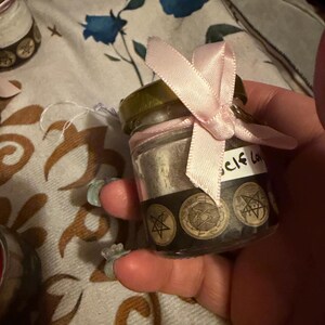 May include: A small clear glass jar with a gold lid, tied with a light pink satin ribbon. The jar contains a light-coloured powder and features a black label around the base with repeating pentagram symbols and the handwritten text Self Love.
