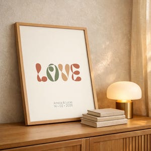May include: A framed print with the word "LOVE" in a retro font, featuring shades of green, orange, and beige. The print includes the names "Amelia & Lucas" and the date "14.02.2026". The print is displayed on a wooden surface.