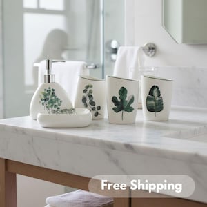 Puede incluir: Un juego de baño de cerámica blanca con diseños botánicos. El juego incluye un dispensador de jabón, un jabonera y dos vasos. Los vasos y el dispensador de jabón tienen ilustraciones de hojas verdes. La jabonera tiene un diseño decorativo. El juego se exhibe sobre una encimera de mármol.