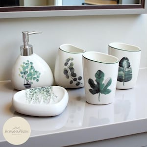 May include: A white ceramic bathroom set with botanical designs. The set includes a soap dispenser, soap dish, and two tumblers, all decorated with green and grey plant illustrations. The dispenser has a silver pump.