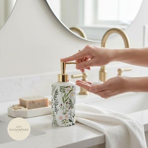 May include: A white ceramic soap dispenser with a gold pump and floral design. The dispenser is being used, dispensing soap into a hand. A bar of soap and a small tray are visible in the background.