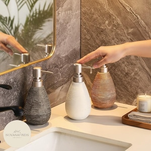 Könnte beinhalten: Ein Set aus drei Keramikbechern mit gesprenkelter, matter Oberfläche. Die Becher sind in den Farbtönen Creme, Hellgrau und Dunkelgrau gehalten. Jeder Becher hat ein schlichtes, minimalistisches Design und einen abgerundeten Henkel.