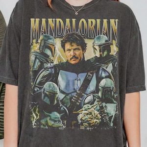 May include: Dark gray t-shirt featuring a graphic of characters from the Mandalorian series. The design includes the word "MANDALORIAN" in gold lettering, along with images of the Mandalorian, Grogu, and other characters in helmets and armor.