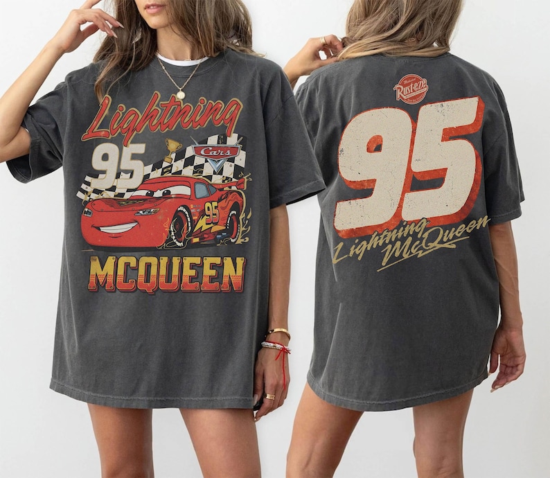 May include: Dark gray t-shirt featuring a graphic of Lightning McQueen from the movie Cars. The front design includes the text "Lightning 95 McQueen" with a race car and checkered flag. The back of the shirt displays a large "95" and "Lightning McQueen" text.
