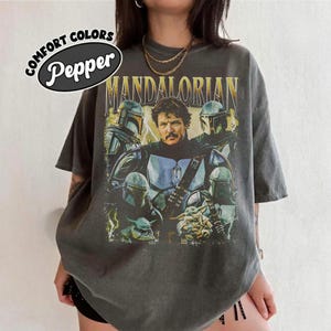 May include: Dark grey t-shirt featuring a graphic of characters from The Mandalorian. The design includes the text "MANDALORIAN" in gold lettering, along with images of the Mandalorian, Grogu, and other characters. The shirt also has the text "COMFORT COLORS Pepper" in a white oval.