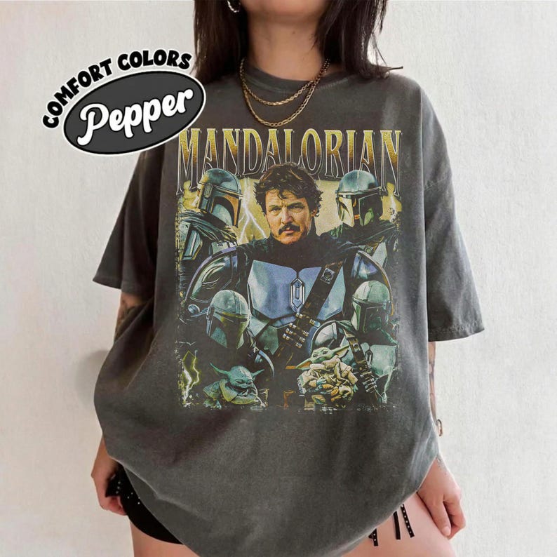 May include: Dark grey t-shirt featuring a graphic of characters from The Mandalorian. The design includes the text "MANDALORIAN" in gold lettering, along with images of the Mandalorian, Grogu, and other characters. The shirt also has the text "COMFORT COLORS Pepper" in a white oval.
