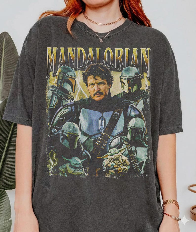 May include: Dark gray t-shirt featuring a graphic of characters from the Mandalorian series. The design includes the word "MANDALORIAN" in gold lettering, along with images of the Mandalorian, Grogu, and other characters in helmets and armor.