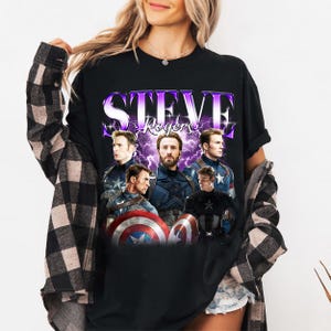 May include: Black t-shirt featuring a graphic of Steve Rogers with the text "STEVE Rogers" in a purple and white design. The graphic includes multiple images of the character in his Captain America uniform, with a shield.