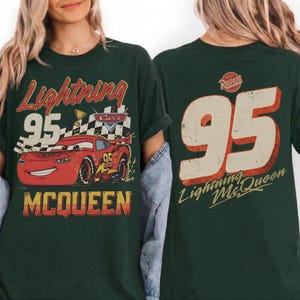 May include: Dark green t-shirt featuring a distressed graphic of Lightning McQueen race car with the number 95. The front of the shirt has the text "Lightning McQueen" and the back has the number 95 and "Lightning McQueen".