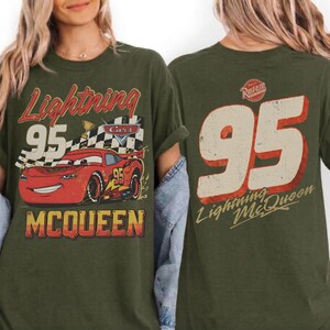 May include: Olive green t-shirt featuring a distressed graphic of Lightning McQueen from the movie Cars. The front design includes the number 95, a checkered flag, and the name "McQueen." The back of the shirt displays a large number 95 and the text "Lightning McQueen."