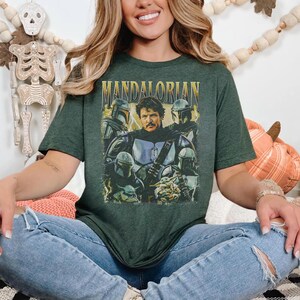 May include: Dark green t-shirt featuring a graphic of the Mandalorian and other characters from the Star Wars series. The word "MANDALORIAN" is printed in gold letters across the top. The shirt is worn by a person wearing blue jeans.