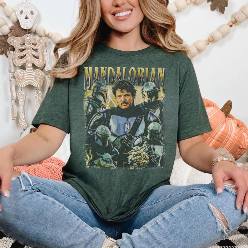 May include: Dark green t-shirt featuring a graphic of the Mandalorian and other characters from the Star Wars series. The word "MANDALORIAN" is printed in gold letters across the top. The shirt is worn by a person wearing blue jeans.