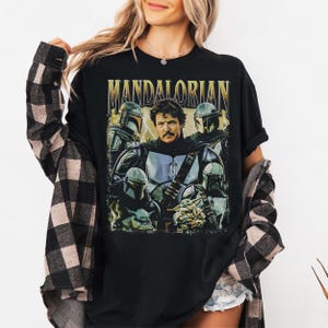 May include: Black t-shirt featuring a graphic of the Mandalorian and other characters from the Star Wars series. The word "MANDALORIAN" is printed across the top in gold lettering. The design includes images of the Mandalorian, Grogu, and other characters.