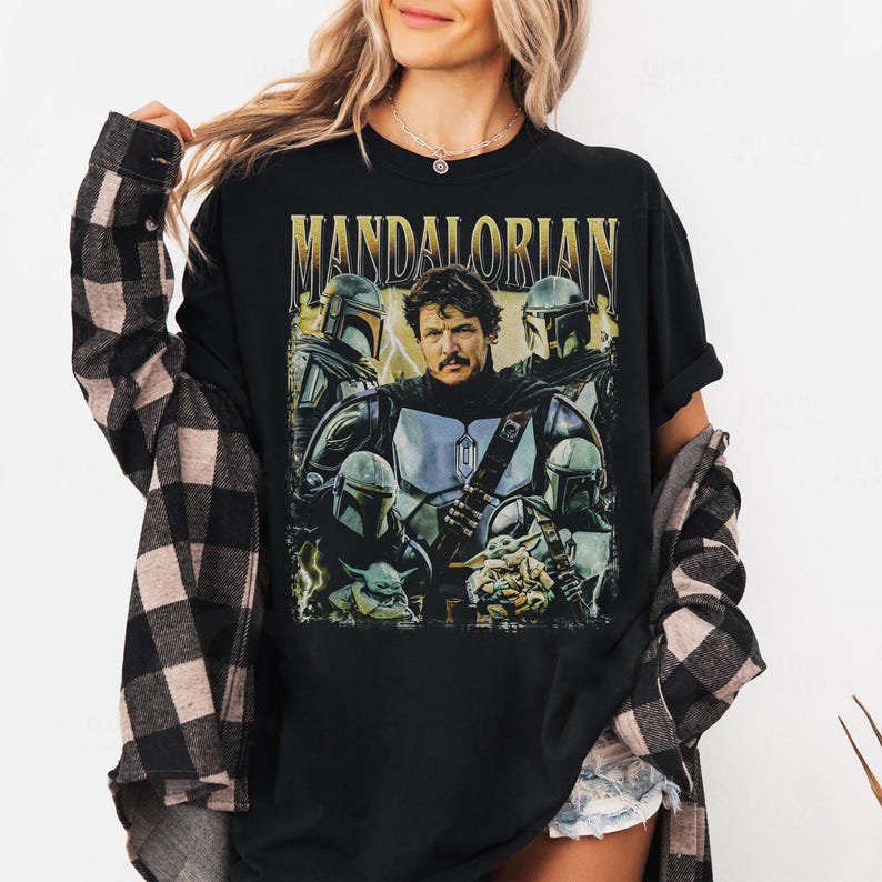 May include: Black t-shirt featuring a graphic of the Mandalorian and other characters from the Star Wars series. The word "MANDALORIAN" is printed across the top in gold lettering. The design includes images of the Mandalorian, Grogu, and other characters.