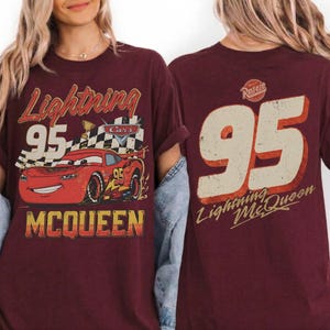May include: Burgundy t-shirt featuring a graphic of Lightning McQueen from the Cars movie. The front design includes the text "Lightning 95 McQueen" with a checkered flag and a smiling red race car. The back of the shirt displays a large "95" and "Lightning McQueen".