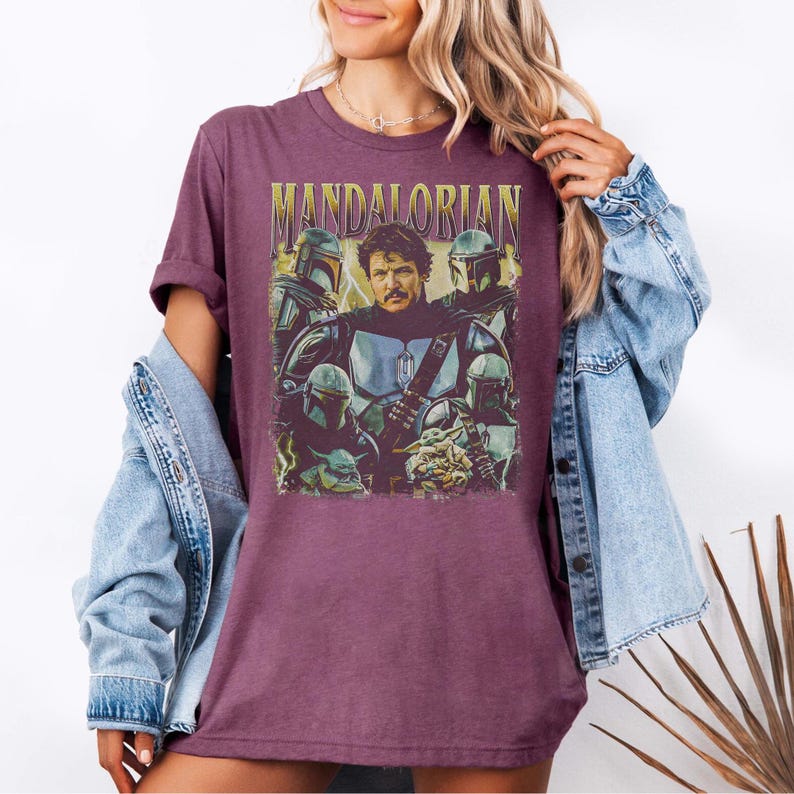 May include: A maroon t-shirt featuring a graphic of characters from the Mandalorian series. The design includes the text "MANDALORIAN" in gold lettering above a group of characters, including the Mandalorian and Grogu. The shirt is worn with a denim jacket.