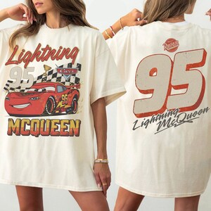May include: Cream-colored t-shirt featuring a graphic of Lightning McQueen from the movie Cars. The front displays the car, the number 95, and the text "Lightning McQueen". The back shows a large number 95 and the text "Lightning McQueen".