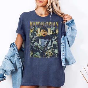 May include: Navy blue t-shirt featuring a graphic of the Mandalorian and other characters from the Star Wars series. The word "MANDALORIAN" is printed across the top in gold lettering. The shirt is a comfortable, casual style.